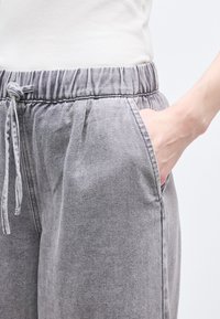 Άτομο που φοράει ανοιχτό γκρι παντελόνι ντένιμ με ελαστική μέση και κορδόνι, λευκό τοπ, χέρι ακουμπισμένο στην μπροστινή τσέπη, ουδέτερο φόντο.