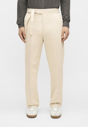 Mężczyzna noszący beżowe, wysokie w talii, szerokie spodnie z materiałowym paskiem, w połączeniu z białymi trampkami i ciemną koszulą z długim rękawem.