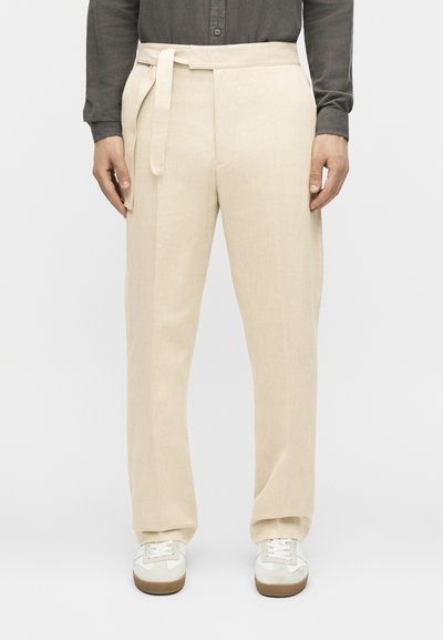 Homme portant un pantalon beige taille haute à jambes larges avec une ceinture en tissu, assorti à des baskets blanches et une chemise sombre à manches longues.