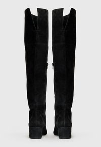 Botas negras de ante, por encima de la rodilla, con un tacón cuadrado, que presentan un diseño con corte en la parte superior y cremalleras laterales para el cierre.