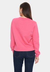 Felpa rosa con maniche lunghe, polsini a coste e scollo rotondo, abbinata a jeans in denim blu scuro visibili nella parte inferiore dell'immagine.