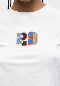 Κοντινό πλάνο ενός ατόμου που φοράει ένα λευκό πουκάμισο με πολύχρωμο κεντημένο αριθμό "23" σε μπλε, μαύρο, πορτοκαλί και κίτρινο στο στήθος.