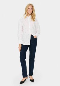 Witte button-up shirt met lichte borduursels, gecombineerd met donkerblauwe jeans. Het shirt heeft een kraag en lange mouwen, met een relaxte pasvorm.
