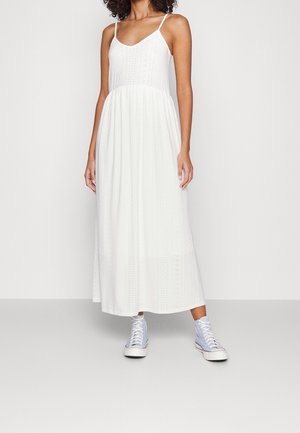 Femme portant une robe maxi blanche sans manches avec des détails en broderie anglaise et des baskets montantes violet clair, debout devant un fond uni.