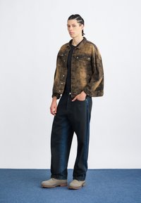 Jeans jakna v rjavi barvi s teksturiranim vzorcem, v kombinaciji s prostornimi črnimi kavbojkami in sivimi škornji z grobim podplatom. Stoji na modri podlagi.