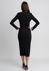 Czarna, dopasowana sukienka midi z długimi rękawami, gładką fakturą i wysokim dekoltem. Posiada subtelne rozcięcie u dołu i jest zestawiona z czarnymi szpilkami.