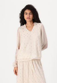 Top beige chiaro con scollatura a V e colletto arricciato, caratterizzato da motivi a pois metallici. Maniche lunghe e trasparenti con polsini elasticizzati. Tessuto morbido e testurizzato.