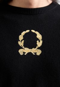 Fekete pulóver bordázott nyakkivágással, kiemelkedő arany hímzett babérkoszorú mintával és a középen található "Moschino" felirattal.