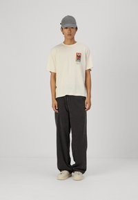 Camiseta crema de manga corta con un gráfico en el pecho, combinada con pantalones de pana negros de corte holgado y una gorra gris. Zapatillas de deporte de color claro.