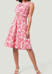 Blumenkleid mit tailliertem Oberteil, ärmellosem Design und weit ausgestelltem Rock. Die Farben umfassen Pink und Weiß mit einem runden Gürtelschnalle-Detail.