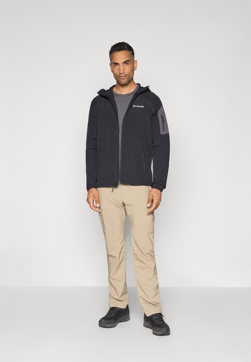 Czarna kurtka zapinana na zamek z kapturem, z szarym akcentem na górnej części ramienia. Nosi się na szarym podkoszulku, w komplecie z beżowymi spodniami i czarnymi butami.