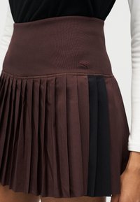 Καφέ πλισέ φούστα με ψηλή μέση και μαύρο πάνελ με μικρό κεντημένο λογότυπο στο δεξί ισχίο, φορεμένη με μαύρο τοπ και λευκό μανίκι που φαίνεται.