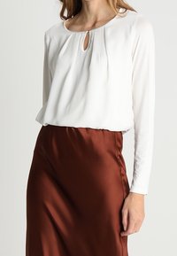 Vit blus med lång ärm och ett nyckelhål i halsringningen, kombinerad med en rostfärgad satinkjol med en slät textur.