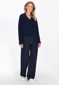 Marineblauwe fuzzy gebreide trui met een V-hals, gecombineerd met wijde, donkerblauwe jeans en witte sneakers. Eenvoudig ontwerp met een ontspannen pasvorm.