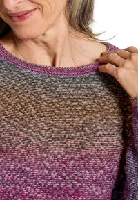 Femme ajustant le col d'un pull tricoté texturé dans des tons de rose et de gris, montrant une partie de son visage et de sa main.