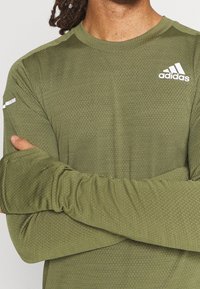 Vihreä oliivinvärinen pitkähihaisen urheilupaita, jossa on tekstuurinen kangas, pyöreä kaula-aukko ja valkoinen Adidas-logo rinnassa ja hihassa.