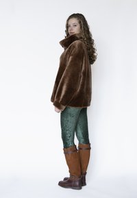 Bruine jasje van nepbont met hoge kraag, gecombineerd met groene getextureerde leggings en bruine leren laarzen tot aan de knie met gespsluitingen.