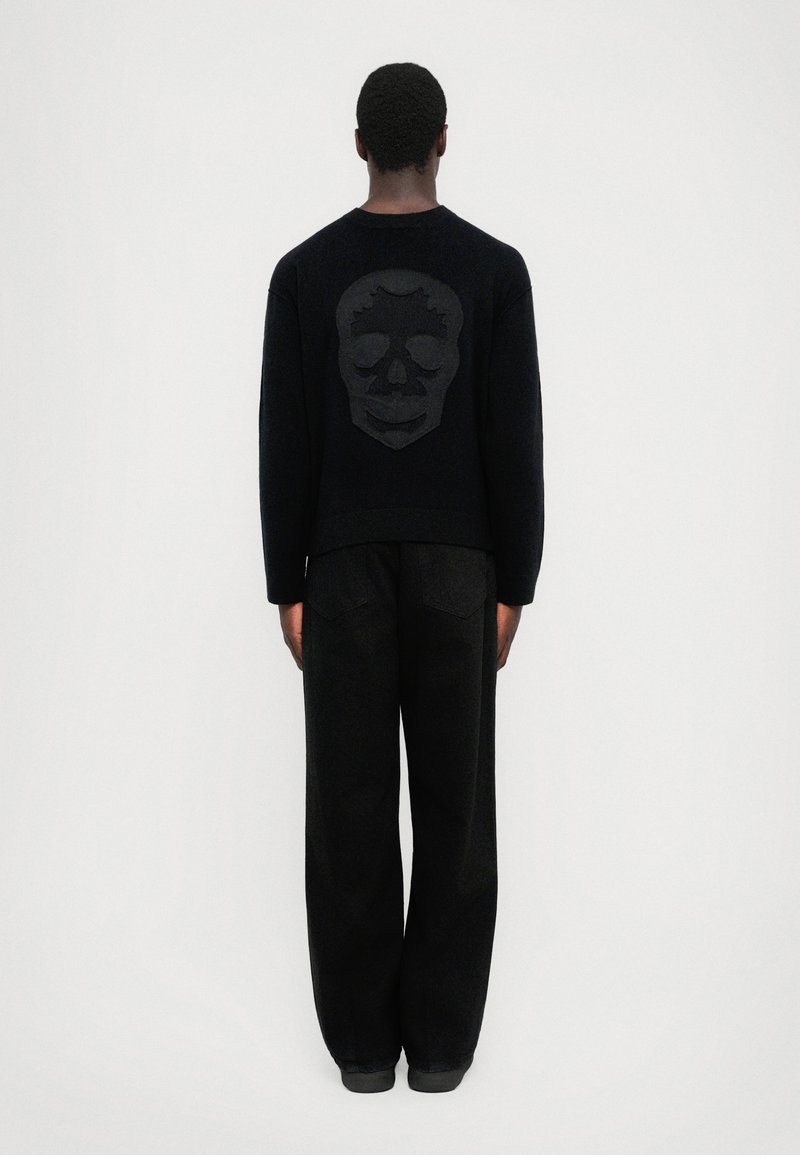 Person facing away wearing black pants and a black sweater with a large textured skull design on the back, standing against white background.