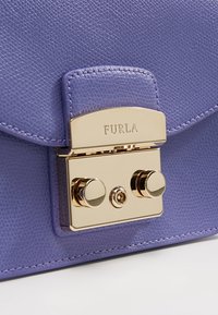 Bolso de cuero texturizado en color púrpura con un cierre dorado que incorpora dos botones redondeados y detalle de marca. Primer plano del diseño frontal.