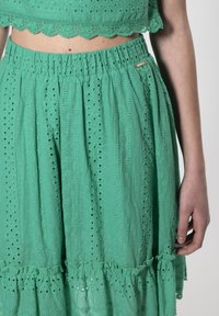 Falda verde en capas hecha de tela ligera con detalles de ojales y un dobladillo con volantes. Cintura elástica para mayor comodidad.