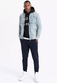 Light blue denim jacket with gray accents, black hooded sweatshirt underneath, navy pants, and white sneakers, standing on a plain background.