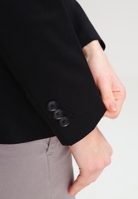 Osoba poprawiająca rękaw czarnej marynarki z trzema czarnymi guzikami, ubrana w jasnoszare spodnie na jednolitym tle.