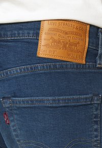 Blå denimjeans med en läderlapp märkt "Levi Strauss & Co." som visar artikelnummer 512, storlek W31 L32, och dekorativ sömnad på bakfickorna.