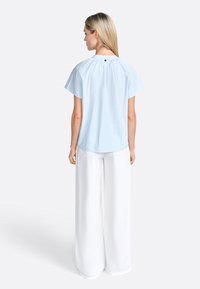 Top a righe blu chiaro con maniche corte e retro plissettato. Abbinato a pantaloni bianchi a gamba larga. Dettaglio dei bottoni evidente sul colletto.