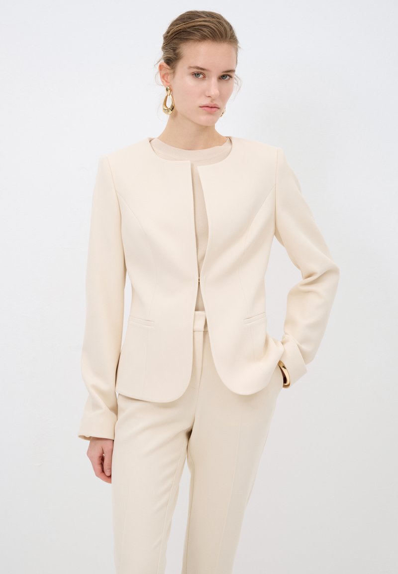 Beige přizpůsobený blazer s kulatým výstřihem, dlouhými rukávy a dvěma předními kapsami. Hladká struktura s přiléhavým siluetem, v kombinaci s odpovídajícími kalhotami.