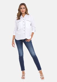 Weißes langärmliges Hemd mit schwarzen Knöpfen und dezenten Seitenstreifen, kombiniert mit dunkelblauen Skinny-Jeans und weißen Riemchensandalen.