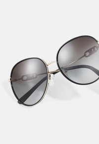 Occhiali da sole oversized neri con lenti grigie sfumate, un accento in metallo dorato sul ponte e punte per le orecchie in plastica. Marchiati con "Michael Kors."
