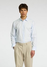 Camicia a righe bleu chiaro con colletto abbottonato e polsini singoli, abbinata a pantaloni beige plissettati, indossata da un modello su uno sfondo neutro.