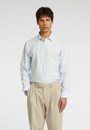 Camicia a righe bleu chiaro con colletto abbottonato e polsini singoli, abbinata a pantaloni beige plissettati, indossata da un modello su uno sfondo neutro.