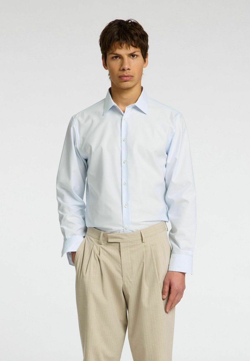 Camicia a righe bleu chiaro con colletto abbottonato e polsini singoli, abbinata a pantaloni beige plissettati, indossata da un modello su uno sfondo neutro.