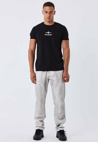T-shirt nera in cotone con logo bianco, abbinata a pantaloni cargo leggeri grigi con texture e cuciture visibili.