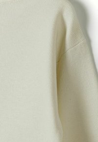 Pull en tricot beige clair avec une texture côtelée, au design simple et aux épaules légèrement imbriquées pour une apparence décontractée.
