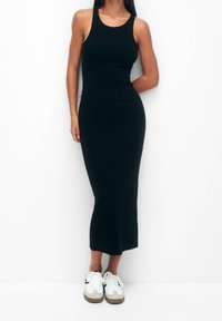 Femme portant une robe midi noire sans manches ajustée associée à des baskets blanches, se tenant devant un fond blanc uni.