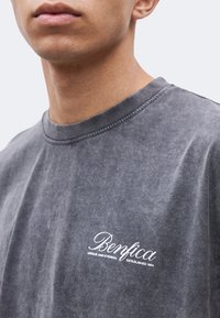 Κοντινό πλάνο ενός ατόμου που φοράει ένα ξεθωριασμένο γκρι μπλουζάκι με το κείμενο «Benfica Unique and Eternal Established 1904» στο στήθος.