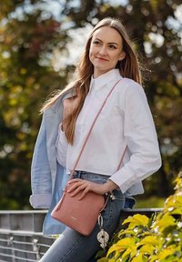 Mladá žena s dlouhými vlasy, oblečená v bílém tričku a džínách, drží růžovou kabelku přes rameno, stojí venku s bundou přehozenou přes rameno.