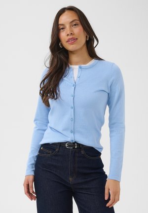 Kvinde med langt mørkt hår iført lyseblå knapforsyet cardigan over hvid top og mørke jeans med sort bælte, stående mod en ensfarvet baggrund.