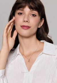 Złoty naszyjnik z wisiorkiem w kształcie półksiężyca noszony na teksturowanej białej koszuli z kołnierzykiem. Tło jest neutralne, skupiające się na biżuterii.