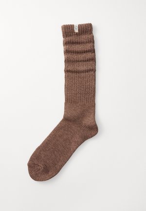 Braune Stricksocke mit geripptem Textur am oberen Ende, glatter Oberfläche am unteren Ende und einem kleinen dekorativen Etikett am Bündchen.