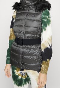 Pufferväst i mörkgrått med hög krage och pälsdetaljer. Har midjebälte och quiltad textur, tillsammans med en långärmad t-shirt i tie-dye.