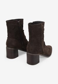 Stivali ankle in suede marrone con un design morbido, dotati di una zip laterale e un tacco largo in legno. Texture liscia con pochi dettagli.
