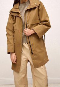 Bruine waterdichte jas met drukknoopsluiting, een hoge kraag en grote voorzakken. Gecombineerd met een gestructureerde trui en lichtbeige broek.