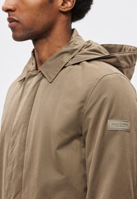 Könnyű barna kabát kapucnival, patentos záródással, oldalzsebekkel és logóval az bal ujján. Simogató tapintású felület.