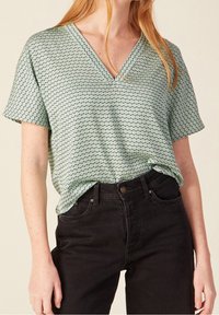 Donna che indossa una blusa a maniche corte con scollo a V a fantasia verde menta, infilata in jeans neri a vita alta, in piedi contro uno sfondo beige.