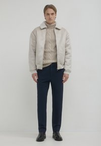 Giacca bomber in shearling beige con colletto di pelliccia bianca, abbinata a un maglione di lana a trecce color crema e pantaloni blu navy. Stivaletti neri alla caviglia completano il look.