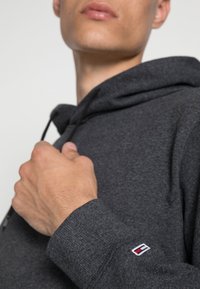 Homme portant un hoodie gris foncé ajustant le col, montrant un gros plan de la main et de la manche avec un petit logo rouge et blanc.