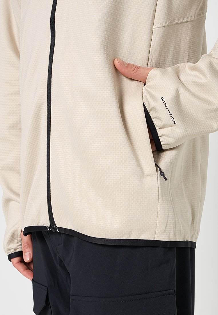 Beżowa kurtka rozpínana wykonana z lekkiego materiału, z czarnymi akcentami, bocznymi kieszeniami oraz subtelnym wzorem fakturowanym na powierzchni.